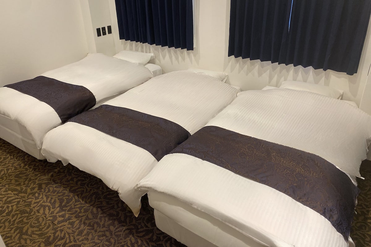 A hotel room with three single beds arranged in a row, each with white bedding and dark brown decorative pillows.
