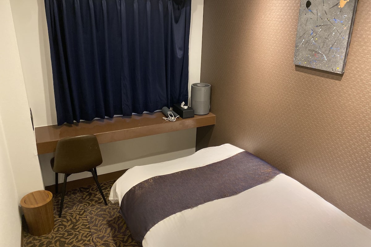 A hotel room with a bed, a desk, a chair, a small trash bin, a window with curtains, and a painting on the wall.
