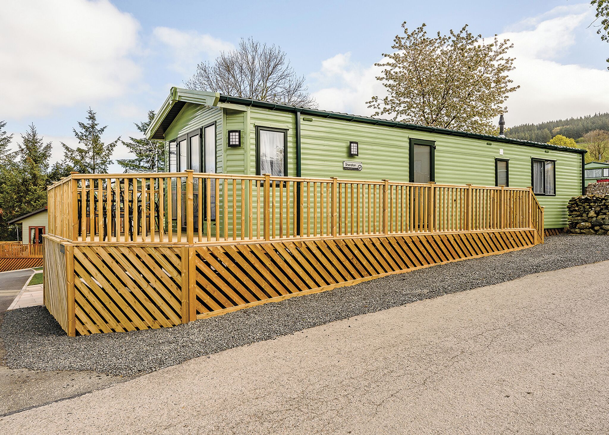 Une maison mobile verte avec une terrasse en bois, une allée en gravier et un ciel dégagé.