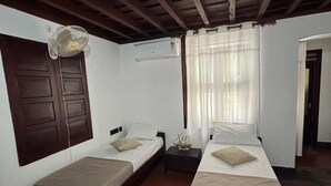 A room with two single beds, a small table, a fan, and a window with curtains. - Vilayara Heritage Retreat (Kozhikode)