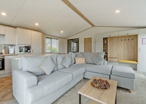 Een moderne woonkamer met een grijze hoekbank, een houten salontafel en een keuken met een magnetron en oven.