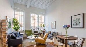 A spacious living room with a sofa, chairs, a coffee table, and a dining table. There are bookshelves, a TV, and artwork on the walls.