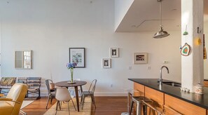 A modern kitchen with a black countertop, stainless steel sink, and bar stools. There is a dining area with a round table and chairs, and a small potted plant.