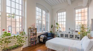 A spacious bedroom with large windows, a bed, a bookshelf, and a potted plant.