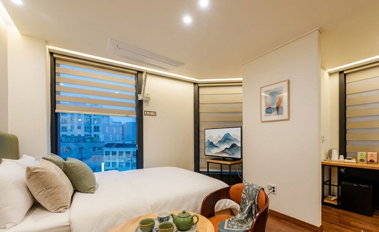 A hotel room with a bed, a table with tea and a teapot, a chair, a desk with a computer, and a painting on the wall.