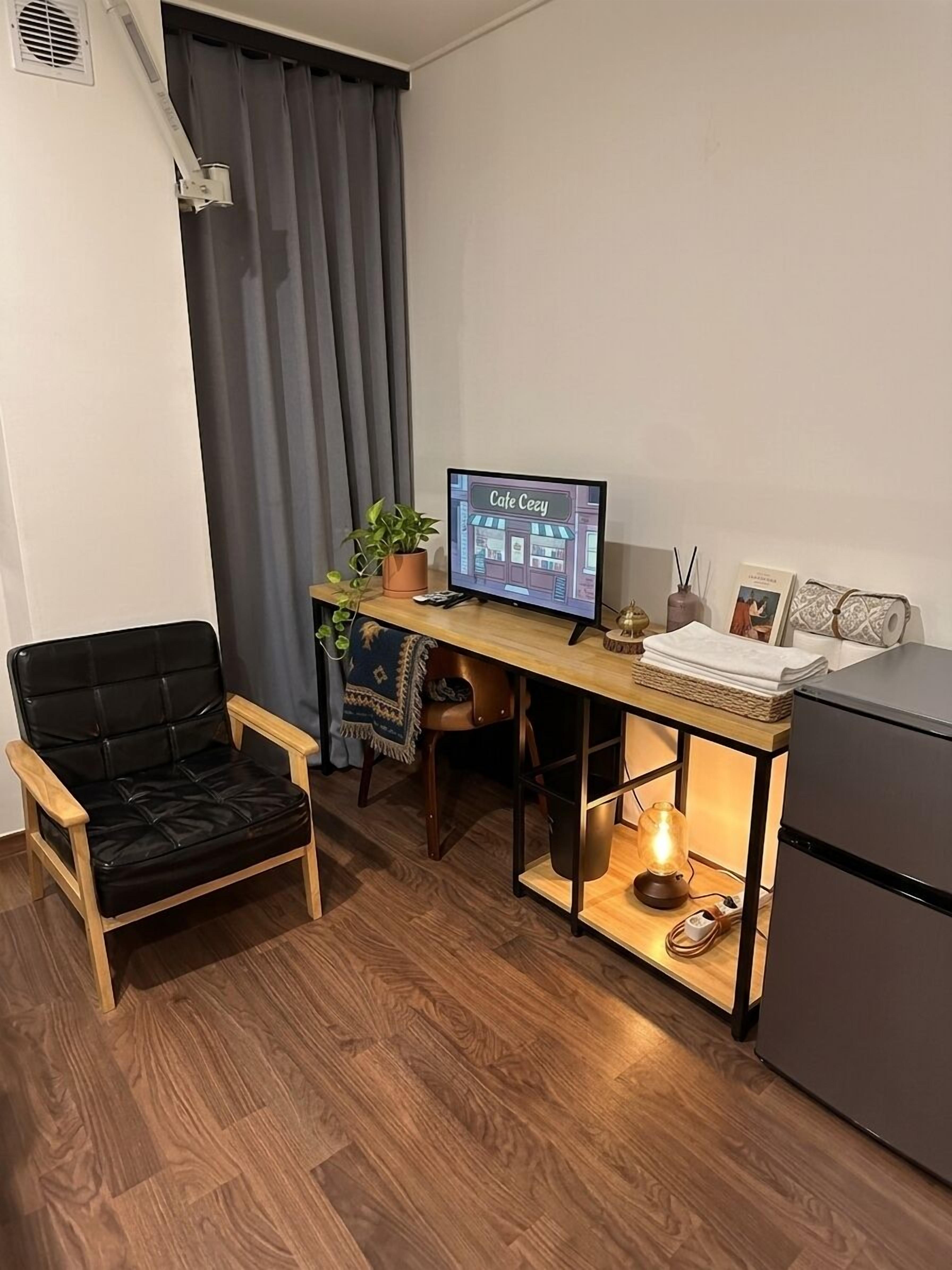 A compact living space with a wooden desk, a black leather chair, a small refrigerator, and a television displaying 'Cafe Cezy'.
