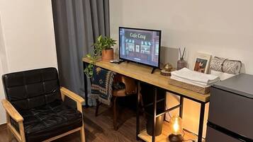 A compact living space with a wooden desk, a black leather chair, a small refrigerator, and a television displaying 'Cafe Cezy'.