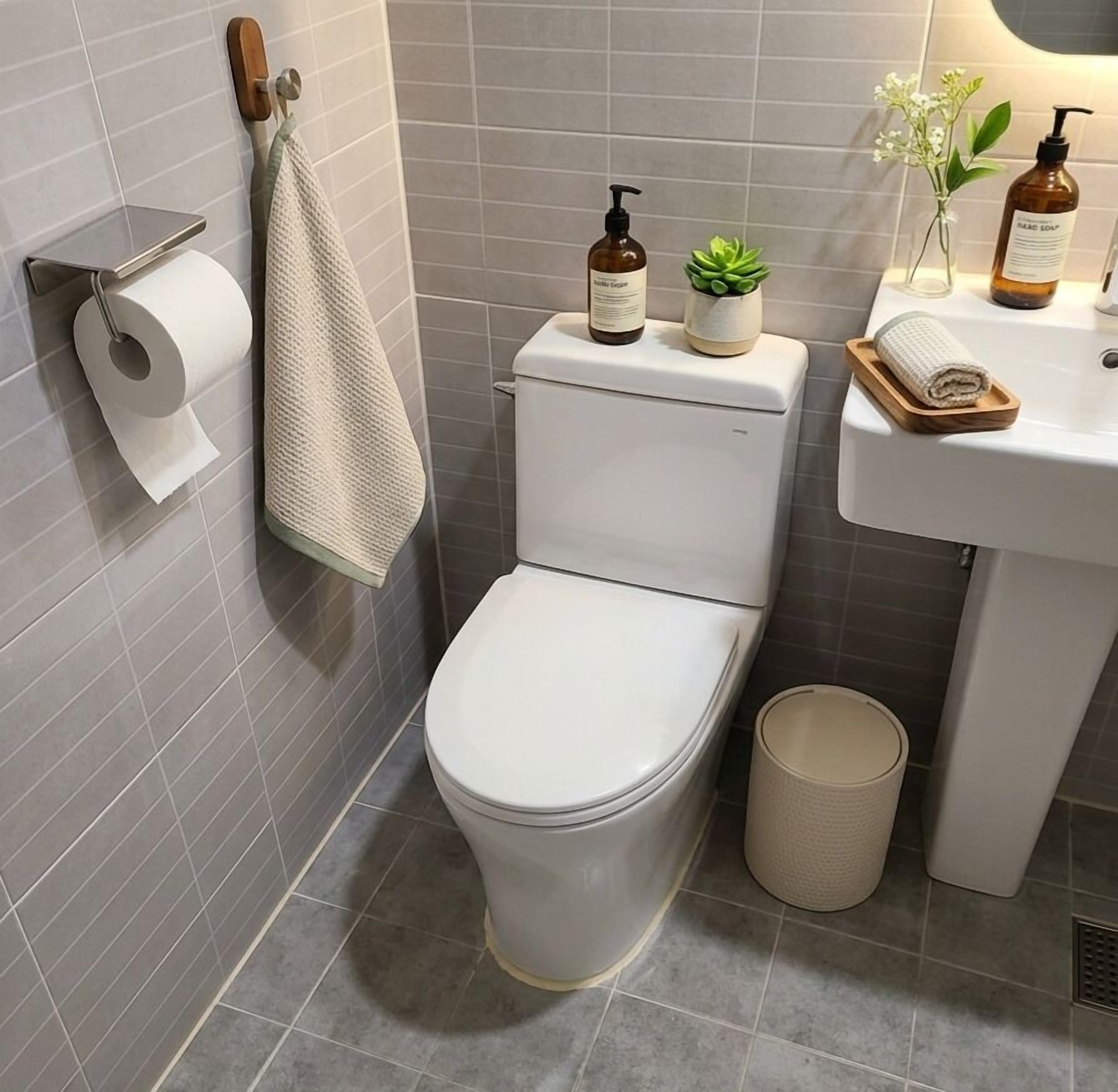 A modern bathroom with a toilet, a sink, a toilet paper holder, a towel, a small plant, and a trash bin.