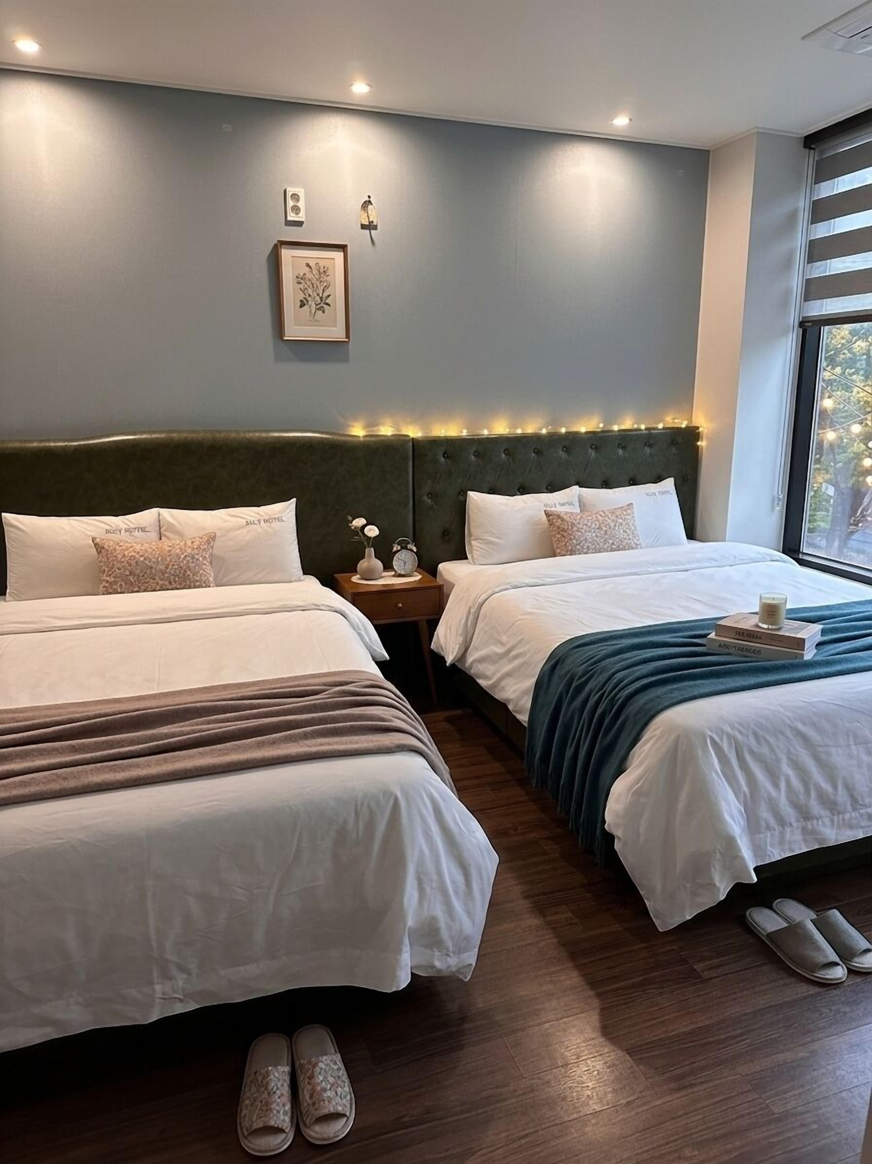 A hotel room with two beds, a nightstand with a clock, a framed picture, and a window with blinds.