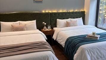 A hotel room with two beds, a nightstand with a clock, a framed picture, and a window with blinds.