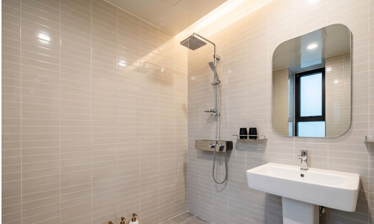 Une salle de bain moderne avec des murs recouverts de carreaux blancs, une douche équipée d’un pommeau de douche à main, un lavabo avec un miroir au-dessus et une petite étagère contenant des articles de toilette.