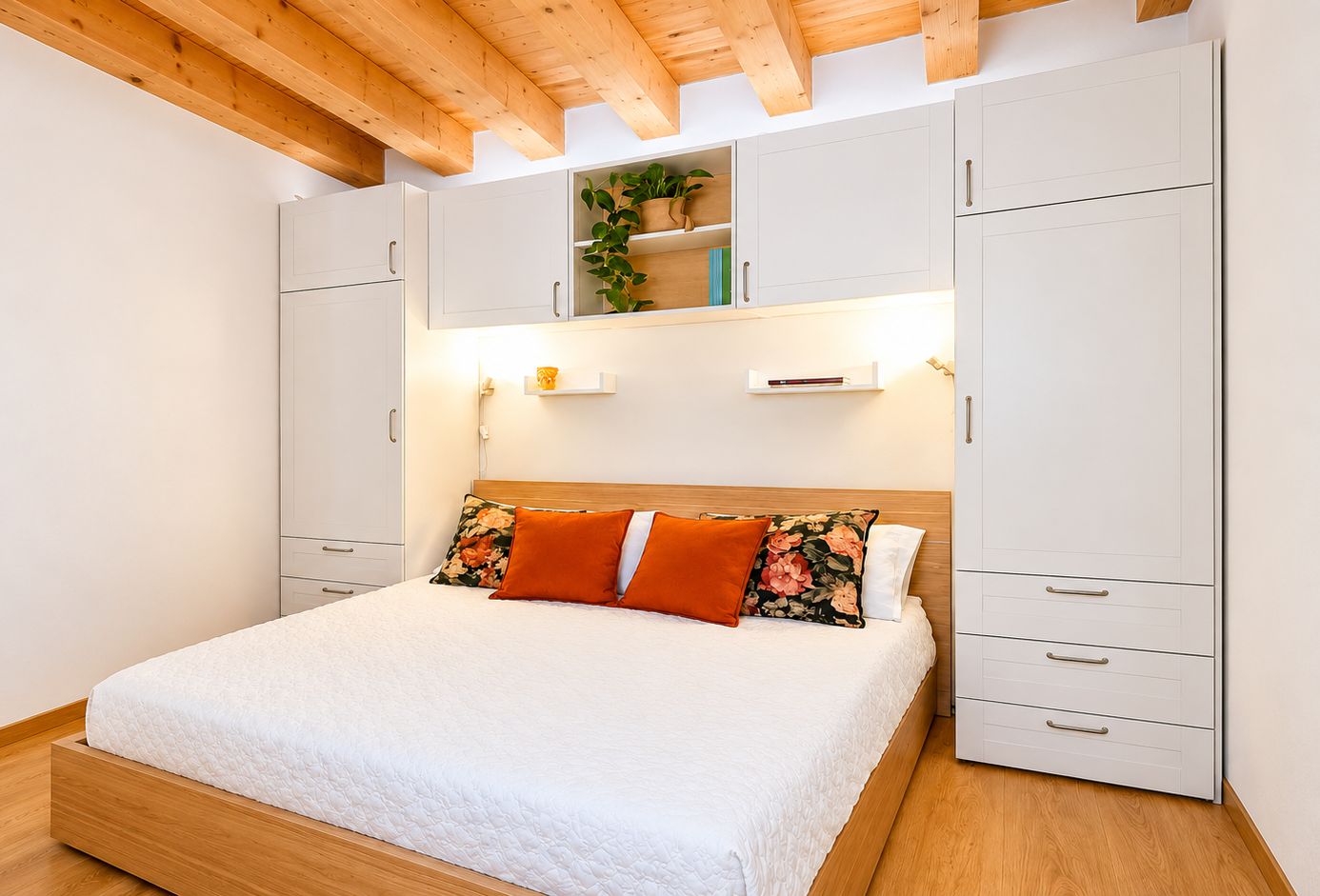 A bedroom with a bed, white cabinets, wooden ceiling beams, and a shelf with plants.