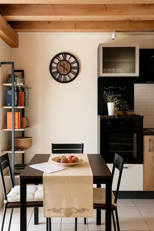 A dining area with a wooden table, chairs, a clock, a bookshelf, and a kitchen in the background.