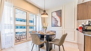 A modern dining area with a round table, four chairs, and a pendant light. A large window with curtains is visible, and a kitchen area with a microwave and kettle is in the background.