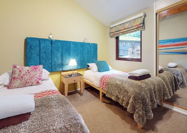 A hotel room with two beds, a bedside table with a lamp, a window with a view, and a mirror.