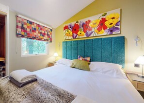 A bedroom with a large bed, a window with a colorful blind, a headboard, a bedside table with a lamp, and a painting of flowers on the wall.