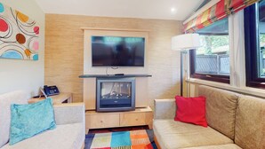 Una sala de estar con una televisión de pantalla plana sobre la chimenea, un sofá con cojines rojos, una alfombra colorida y una ventana con cortinas.