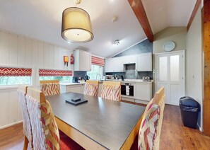 A dining area with a wooden table and patterned chairs, a kitchen with white cabinets and a stove, and a clock on the wall.