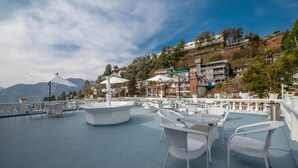 An outdoor terrace with wicker furniture, umbrellas, and a view of a hillside with buildings. - Syona Resort (Mussoorie)
