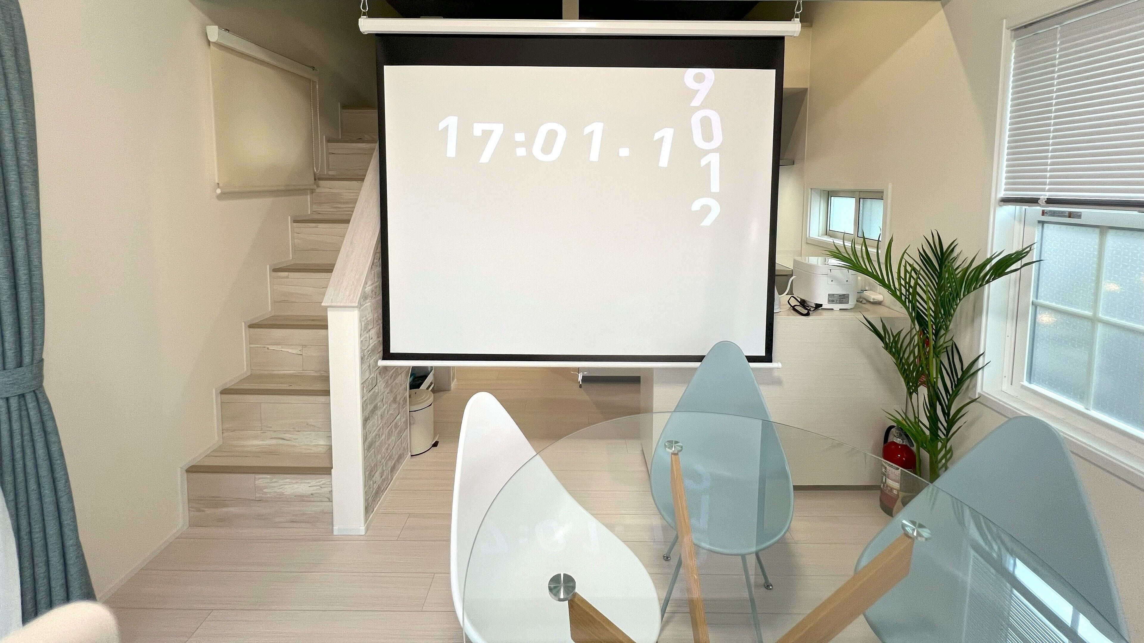 A modern conference room with a projector screen displaying a date and time, a staircase, and a potted plant.