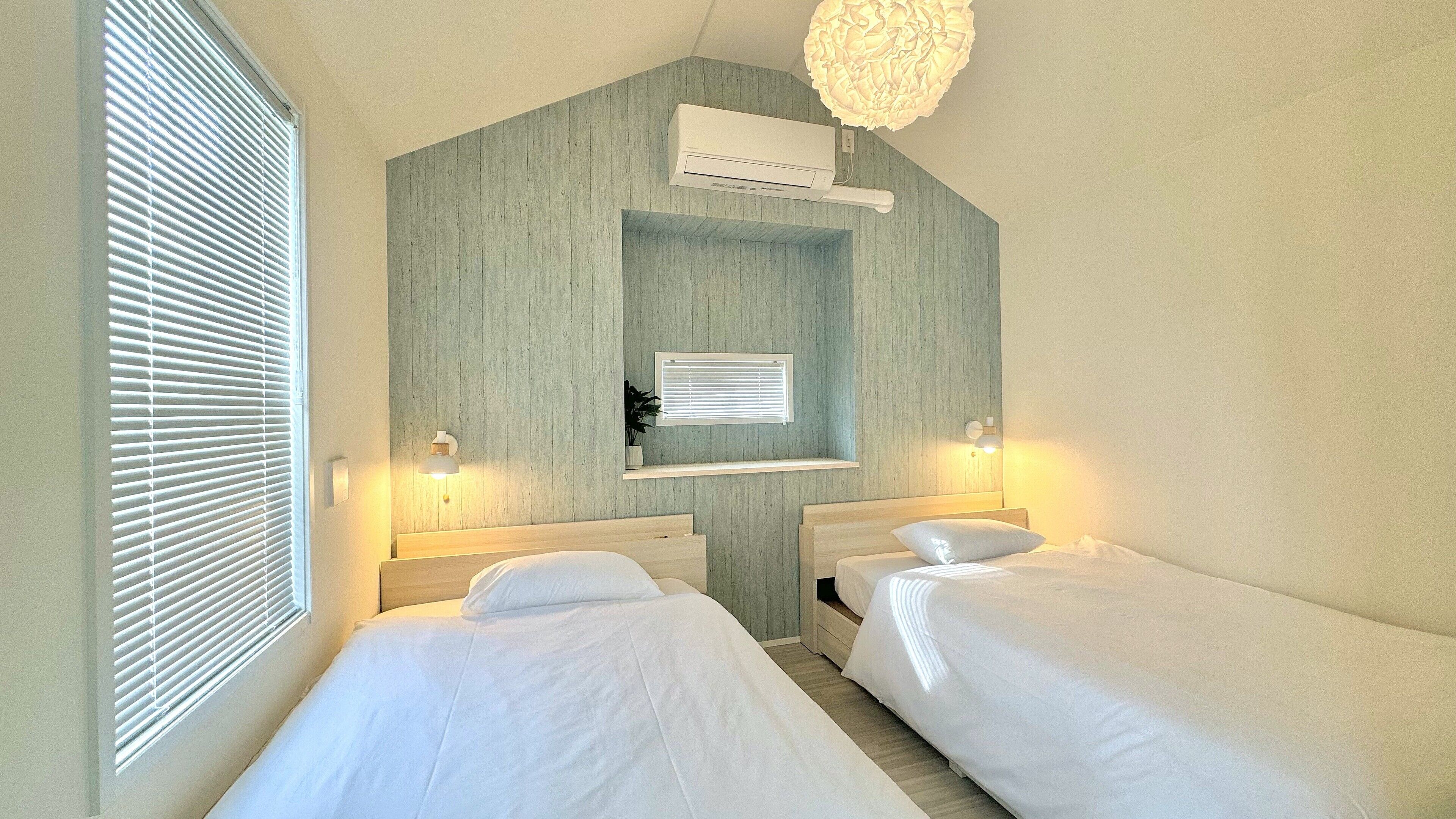 Quarto de hotel com duas camas de solteiro, uma janela com persianas, unidade de ar condicionado, prateleira na parede e luminária no teto.