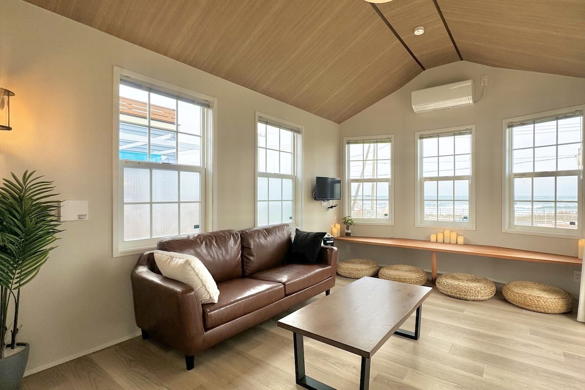 Amplia sala de estar con un sofá de cuero marrón, una mesa de centro de madera y un televisor montado en la pared. Amplias ventanas ofrecen vista a la playa.