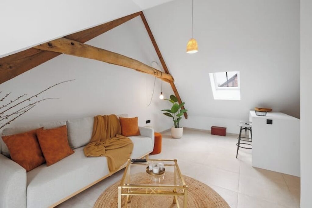 A modern loft apartment with a sloped ceiling, a sofa with cushions, a coffee table, a potted plant, and a small kitchenette.