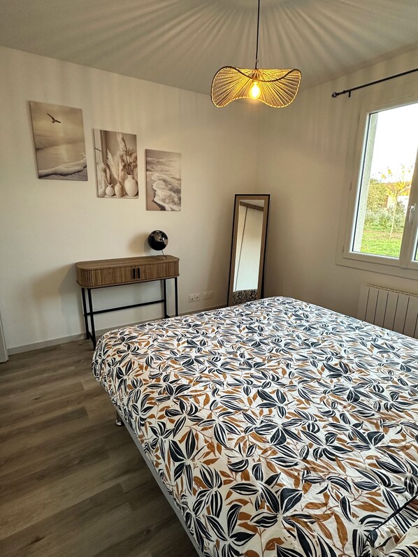A bedroom with a patterned bedspread, a hanging light fixture, a mirror, and wall art.