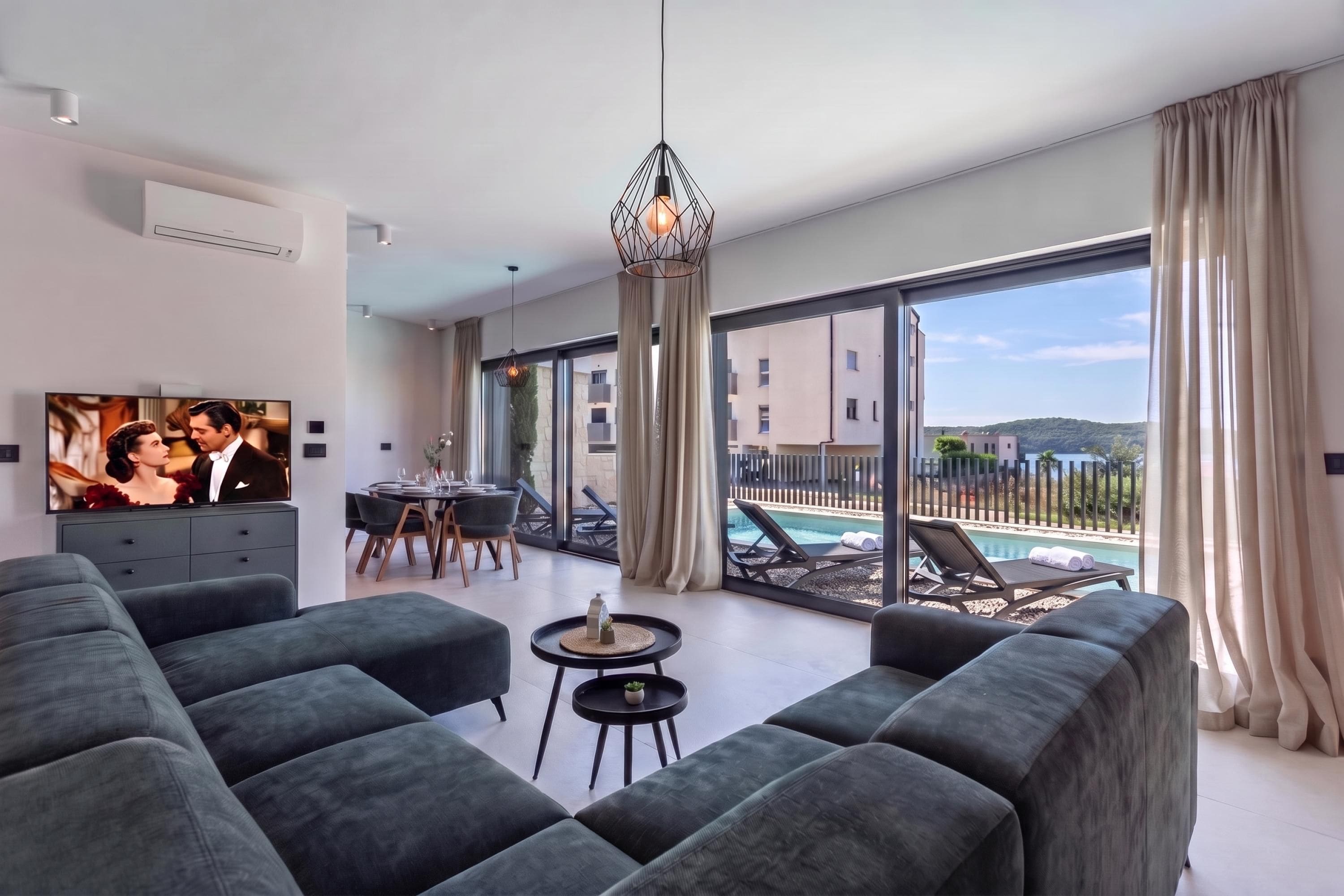 A modern living room with a large flat-screen TV, grey sofas, a dining area with a table and chairs, a hanging light fixture, and a balcony with a pool view.