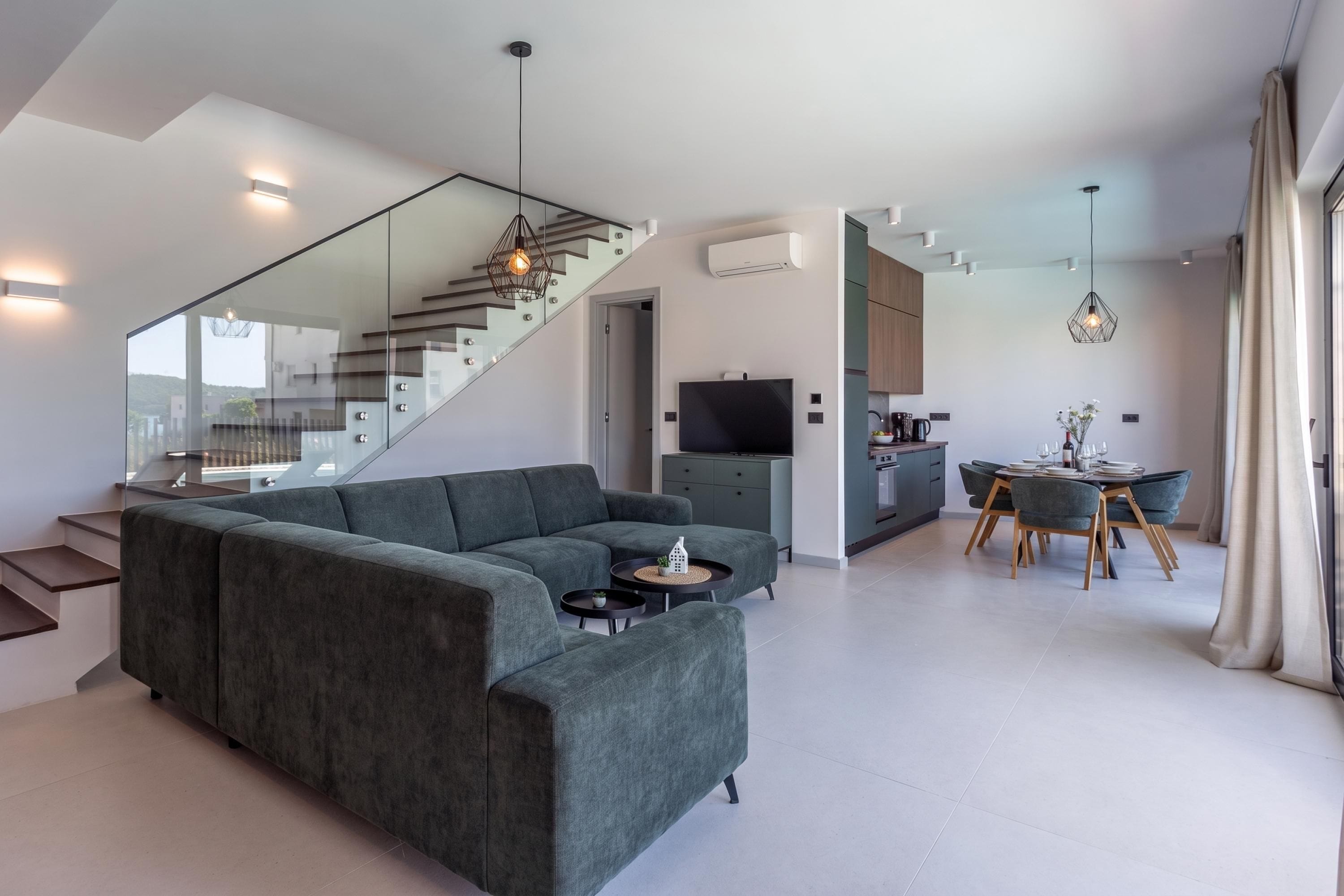 Un saló modern amb un sofà modular, una zona de menjador amb taula i cadires, una cuina amb televisió i una escala amb barana de vidre.