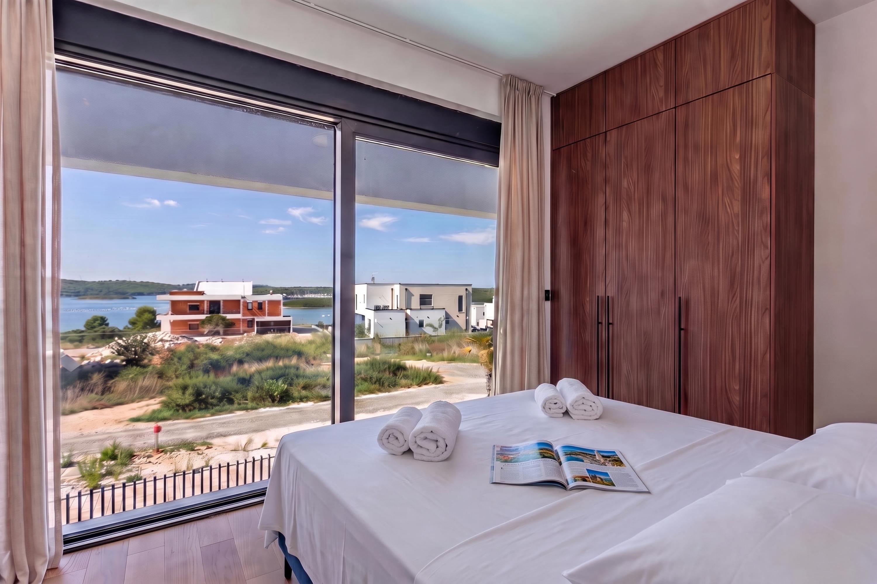 A hotel room with a large window, a bed with white linens, and a view of buildings and greenery.