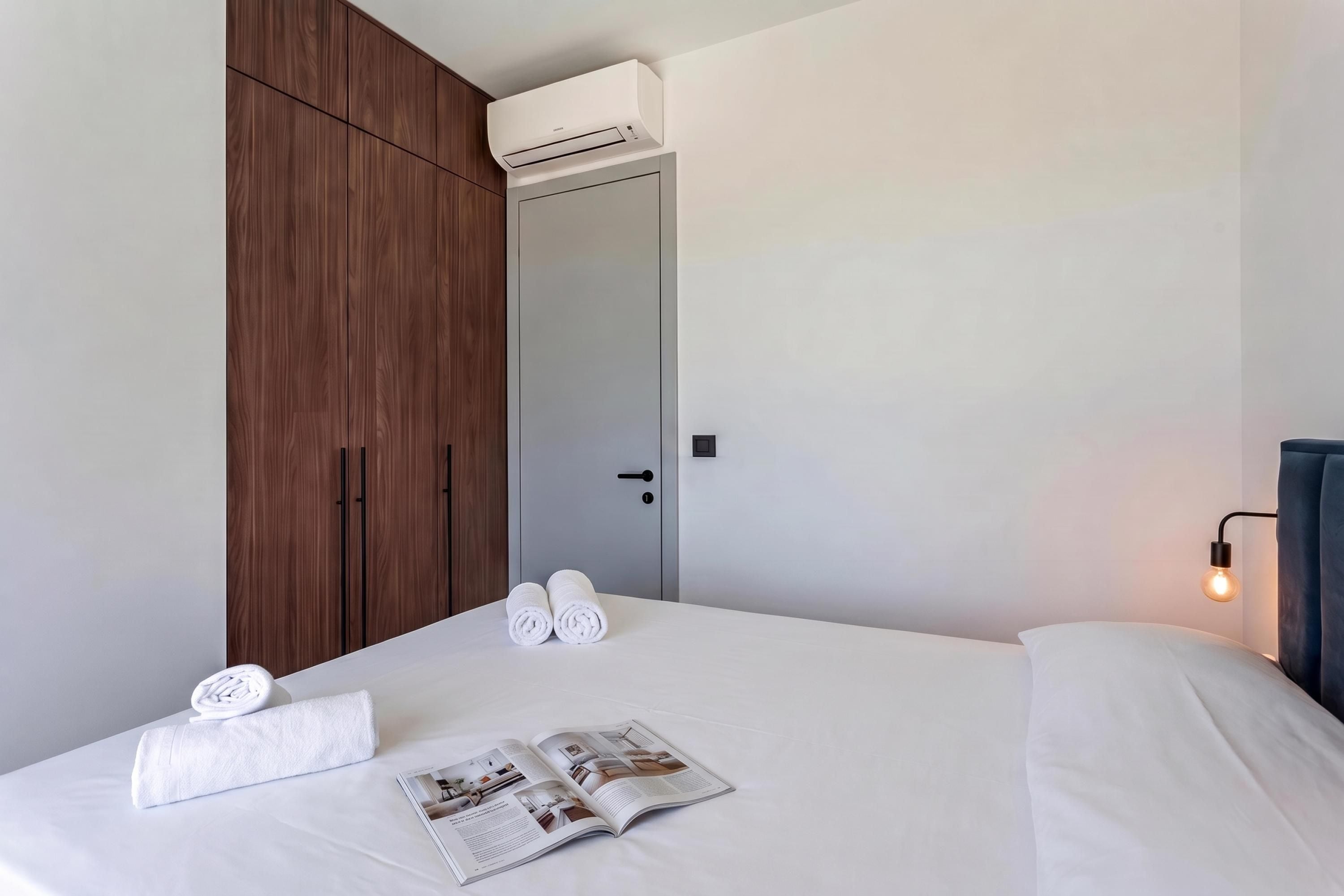 A hotel room with a bed, a wooden wardrobe, an air conditioning unit, a door, a towel, and a magazine.