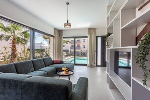 A modern living room with a sectional sofa, a small table with fruit and drinks, a TV on a white shelf, and a view of a swimming pool through large windows.