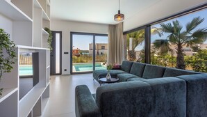 A modern living room with a sectional sofa, a small round table, and a large window offering a view of a swimming pool and palm trees. - Villa Petra with Pool and Sea View (Medulin)