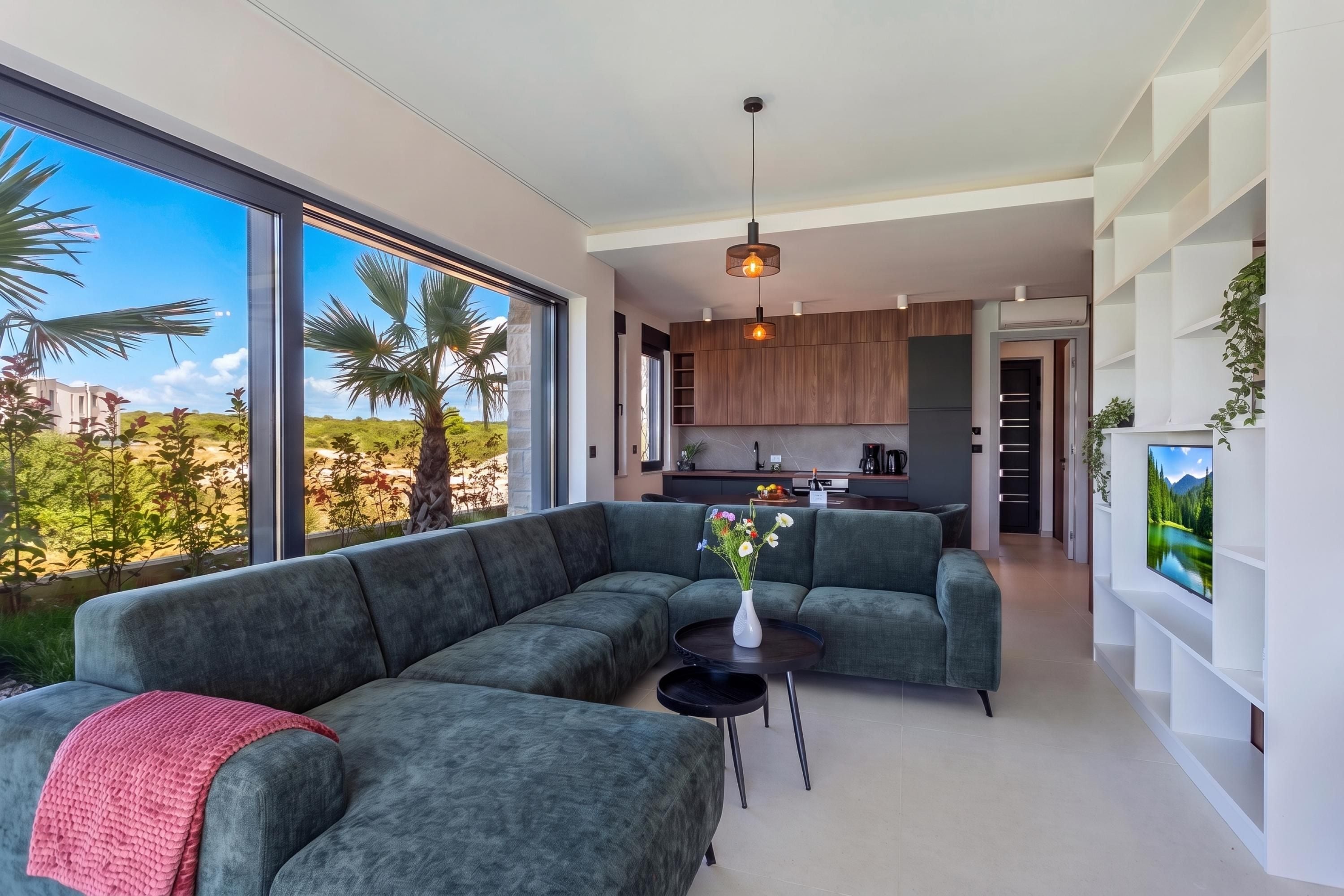 A modern living room with a sectional sofa, a small round table with a vase, and a view of the kitchen and outdoor area.
