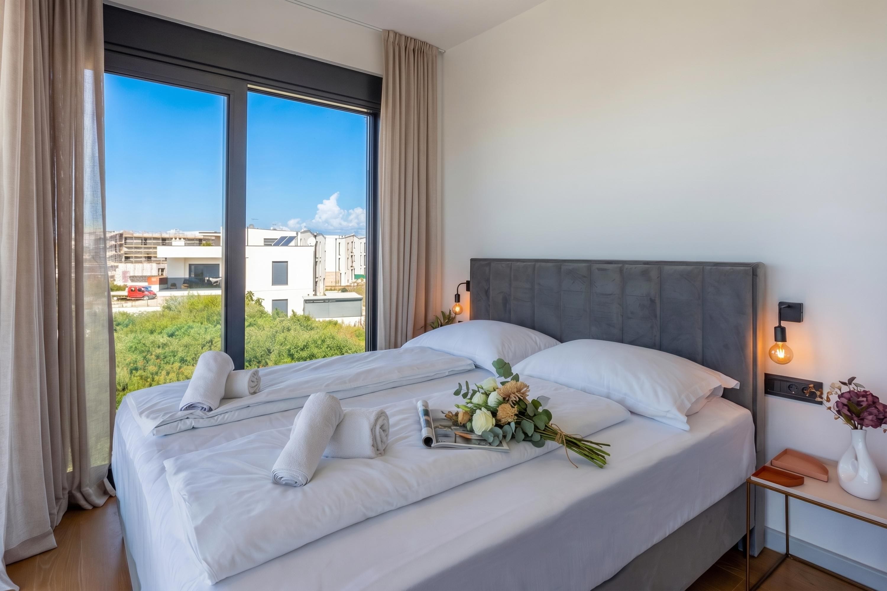 A hotel room with a large bed, a nightstand, a wall-mounted lamp, and a view of buildings through the window.