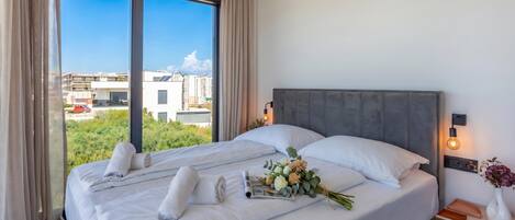 A hotel room with a large bed, a nightstand, a wall-mounted lamp, and a view of buildings through the window.
