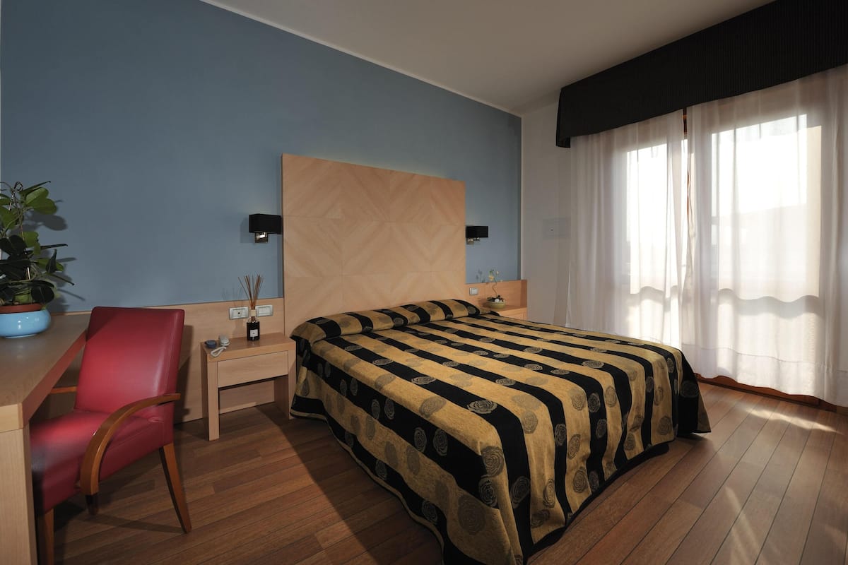 Hotelkamer met een bed, bureau, stoel, nachtkastje, een kamerplant en een raam met gordijnen.