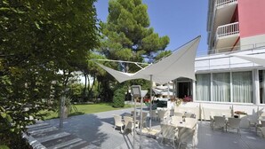 Ένας εξωτερικός χώρος με τραπεζάκια και καρέκλες από λευκό μπαμπού και μια μεγάλη λευκή ομπρέλα που παρέχει σκιά.