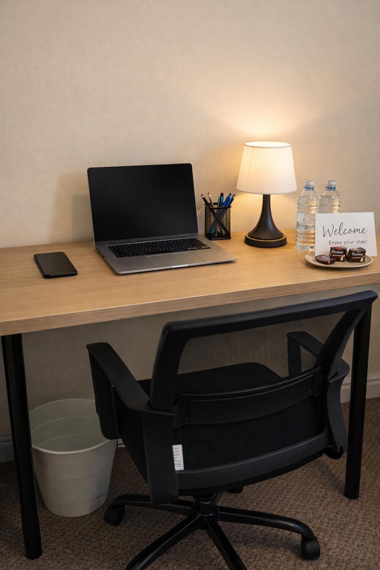 A hotel workspace with a laptop, desk lamp, water bottles, a mobile phone, and a welcome sign.
