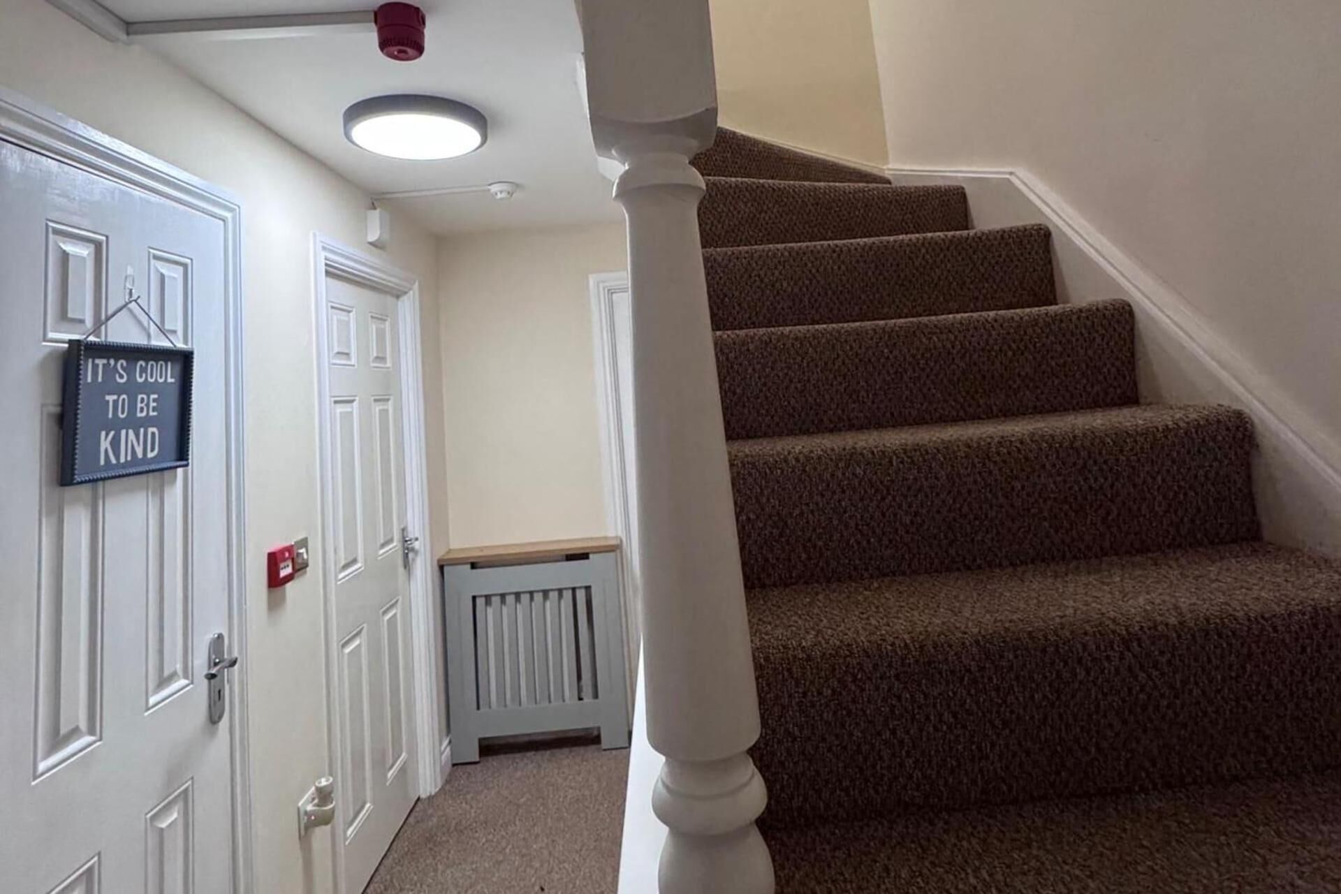 Un escalier dont les marches sont recouvertes de moquette, avec des murs blancs et une porte sur laquelle est apposée une enseigne.