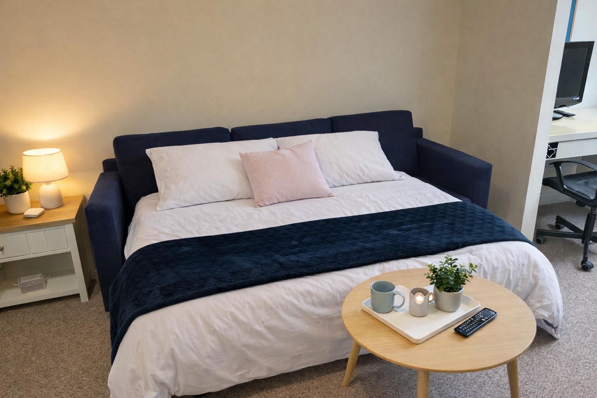 Una cama bien hecha con un cabecero azul oscuro, ropa de cama blanca y una manta azul oscuro. Se ve una mesita de noche con una lámpara, una planta pequeña y un control remoto. Frente a la cama hay una mesa redonda con una bandeja que contiene una taza, un control remoto y una planta pequeña.