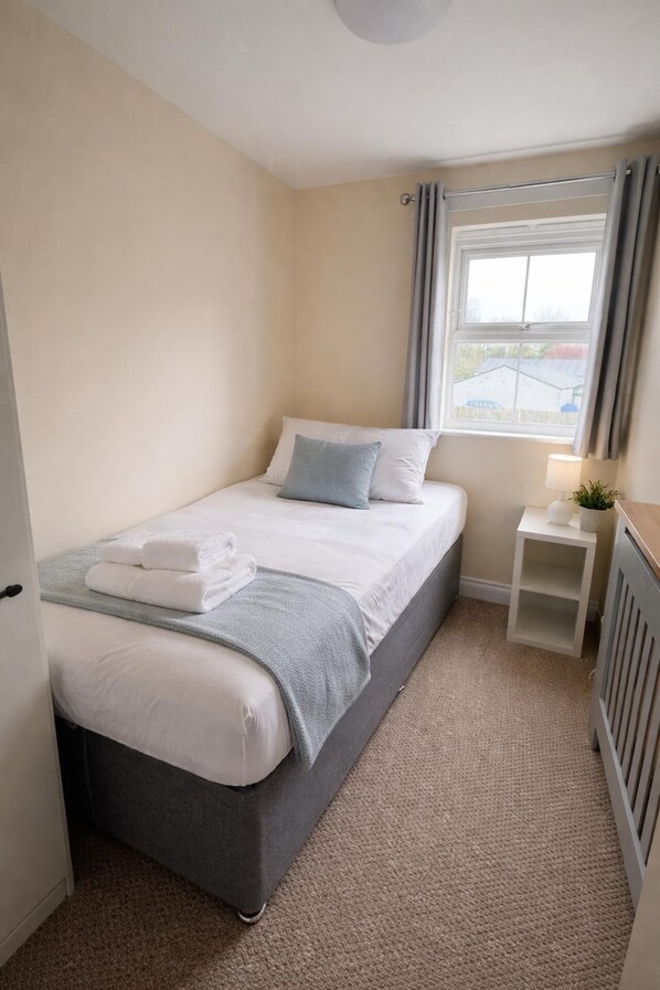 A small, neatly arranged bedroom with a single bed, a bedside table with a lamp, and a window with curtains.