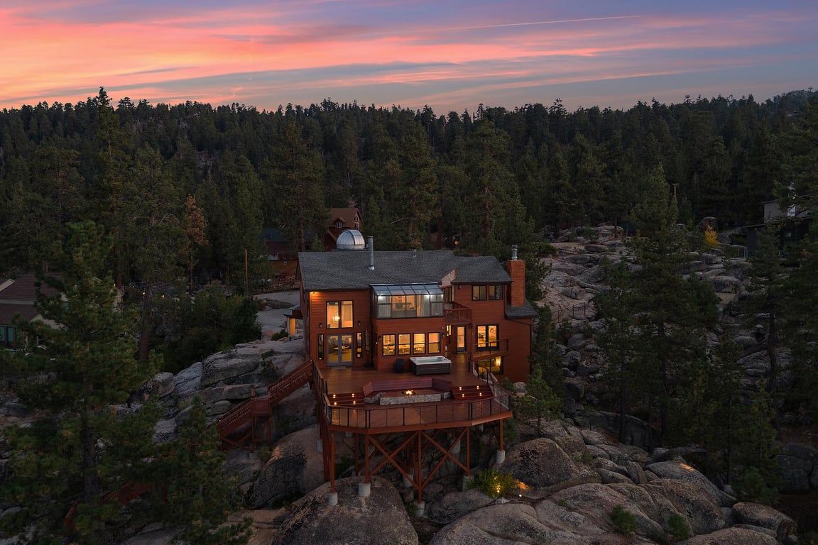 A multi-story wooden house with a deck, surrounded by a forested area.