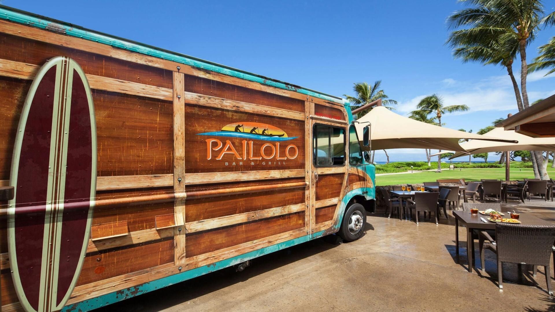 Ein Imbisswagen mit Surfboard-Design, gekennzeichnet als 'Pailolo Bar & Grill', steht draußen mit Tischen und Stühlen unter Sonnenschirmen geparkt.