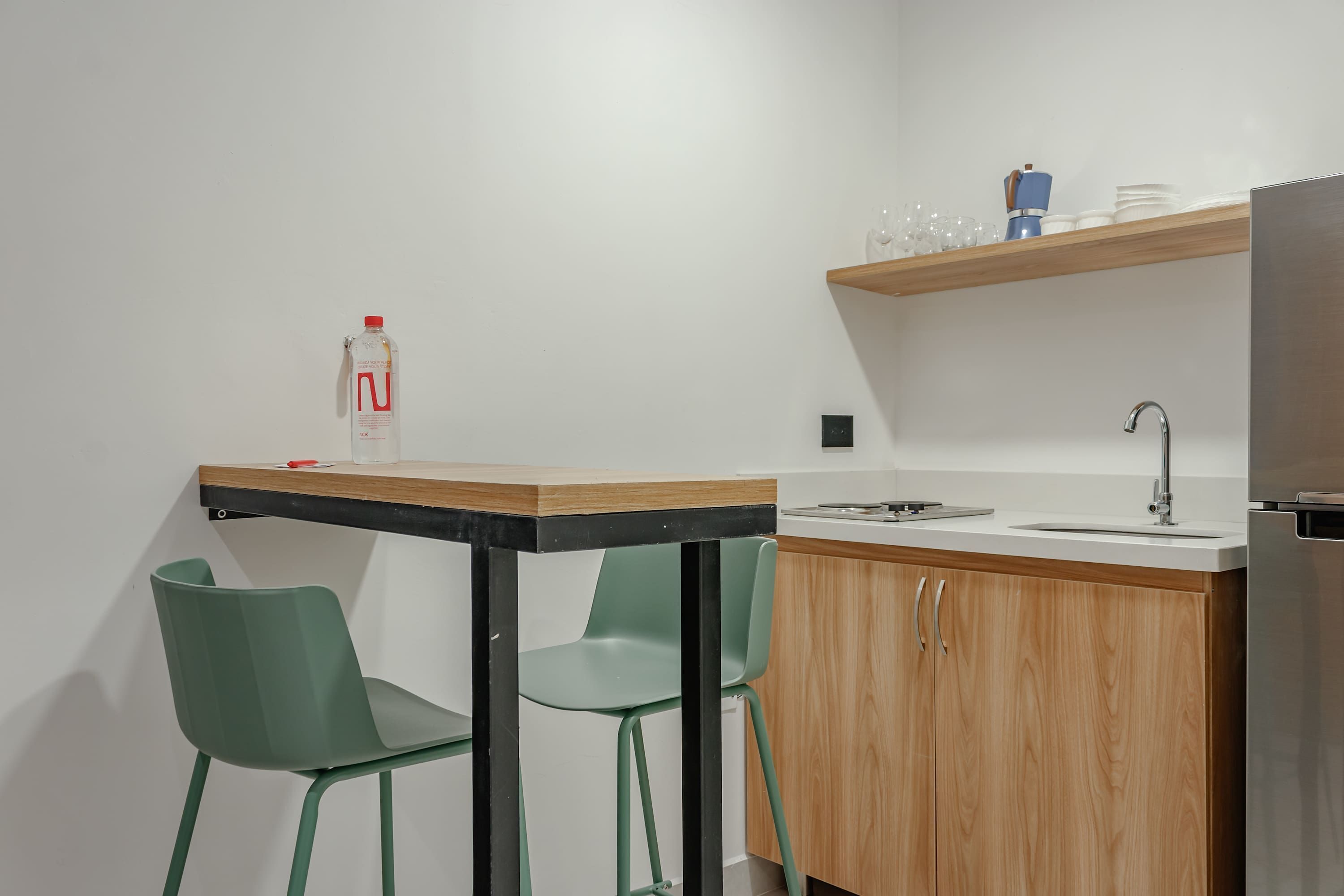 Cozinha compacta com bancada de madeira, pia e geladeira de aço inoxidável. Há duas cadeiras verdes e uma mesa pequena com uma garrafa e um copo.