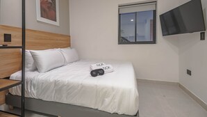 A neatly made bed with white bedding and two rolled towels with text, a wooden headboard, a mounted TV, and a window with a view. - NOK Amazing Loft in Laureles (Medellín)