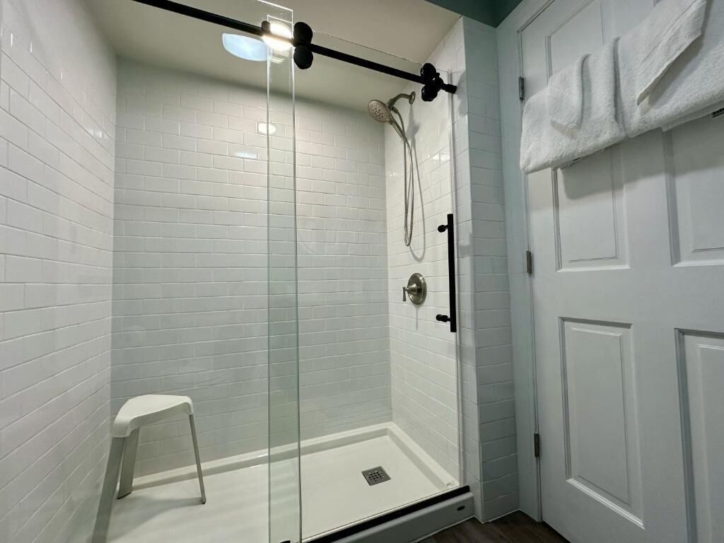 Eine begehbare Dusche mit Glastrennwand, einer weißen Fliesenwand, einer weißen Tür mit einem Handtuch und einem kleinen weißen Hocker.