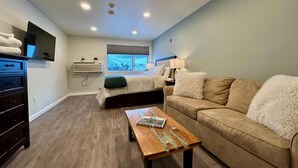 A modern bedroom with a bed, a sofa, a wooden coffee table, a TV, and a window with a view.