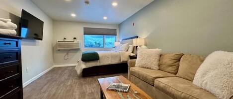 A modern bedroom with a bed, a sofa, a wooden coffee table, a TV, and a window with a view.